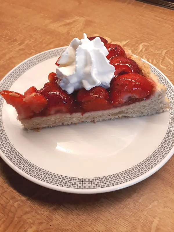 Eine Tortenscheibe mit Erdbeeren und Sahne steht auf einem weißen Teller, umgeben von einer Holzoberfläche.