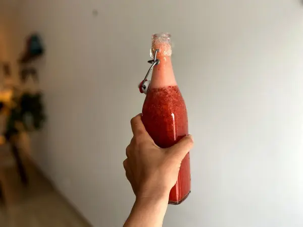 Eine Hand hält eine Flasche mit rosa Erdbeer-Limes, auf einem neutralen Hintergrund, bereit für die Grillparty.
