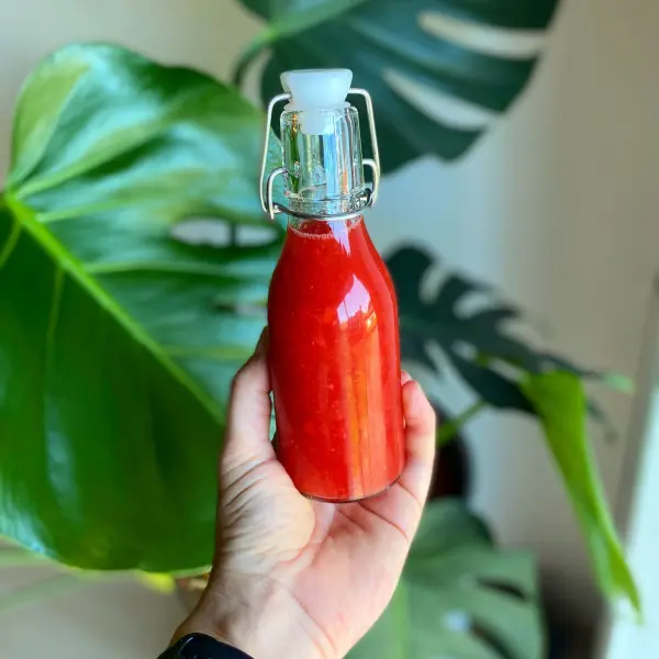 Eine Hand hält eine Flasche mit erdbeerfarbenem Limes vor grünem Blattwerk, ideal für die Grillparty.