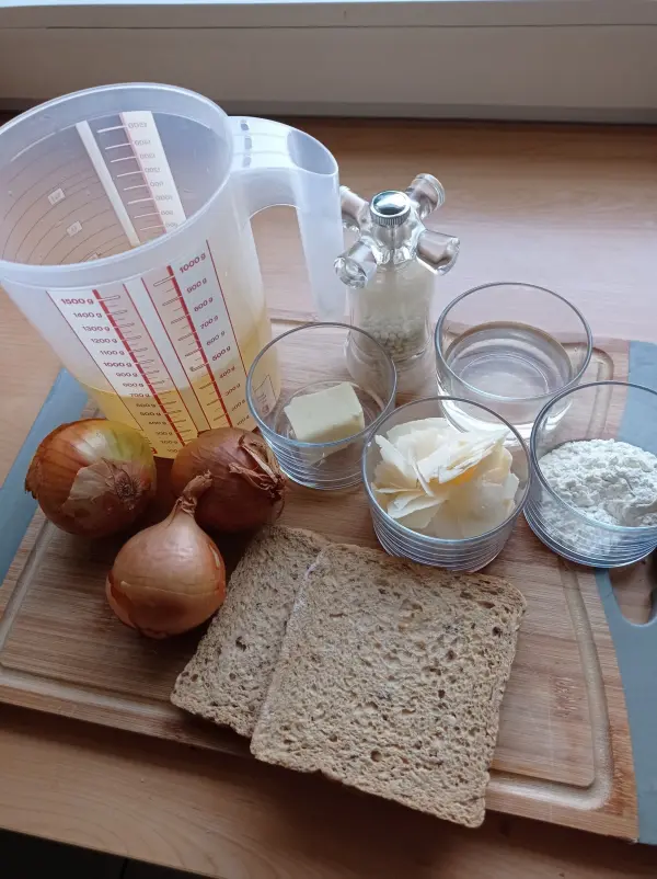 Zwiebeln, Brühe und Zutaten für die französische Zwiebelsuppe sind auf einem Holzbrett in einer Küche angeordnet.