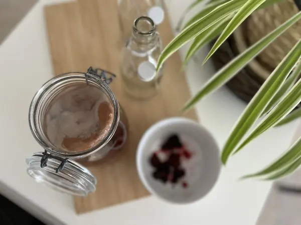 Ein Glas mit fermentiertem Tee steht auf einem Holzteller, umgeben von Flaschen und einer Schale mit Früchten.