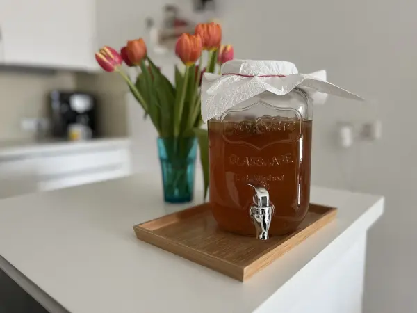Ein Glasbehälter mit Kombucha steht auf einem Holztablett, umgeben von frischen Tulpen in einer Vase.