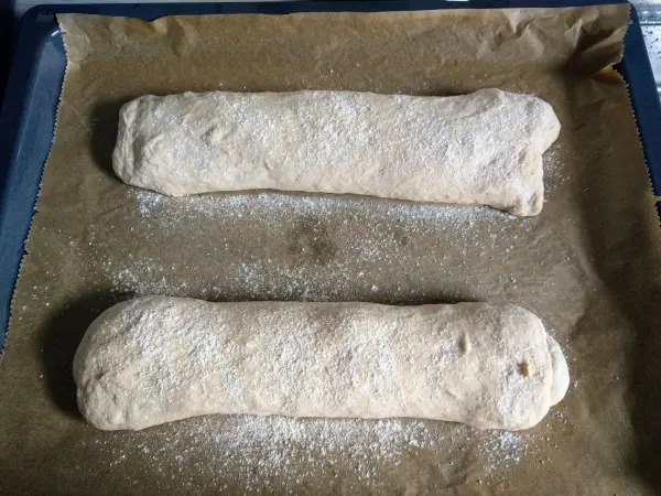 Zwei gestreckte Dinkel-Ciabatta-Rohlinge liegen auf einem Backblech mit Backpapier, leicht mit Mehl bestäubt.