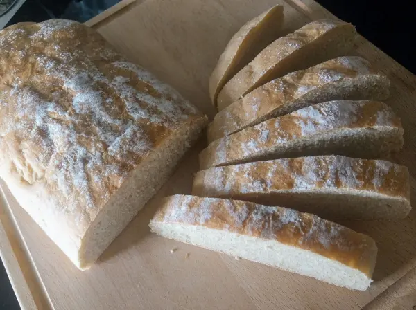 Ein frisch gebackenes Dinkel-Ciabatta liegt auf einem Holzbrett, in gleichmäßige Scheiben geschnitten und leicht bestäubt.