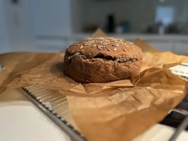 Ein knuspriges Roggenvollkorn-Sauerteigbrot ruht auf einem Backgitter, umgeben von braunem Backpapier, in einer hellen Küche.