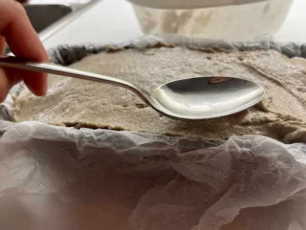 Der silberne Löffel hebt Teig aus einer Backform, um knuspriges Roggenvollkorn-Sauerteigbrot vorzubereiten.