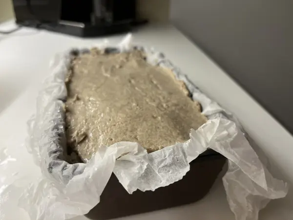Ein Roggenvollkorn-Sauerteigbrot in einer Backform, das auf einer weißen Oberfläche ruht und auf das Backen wartet.