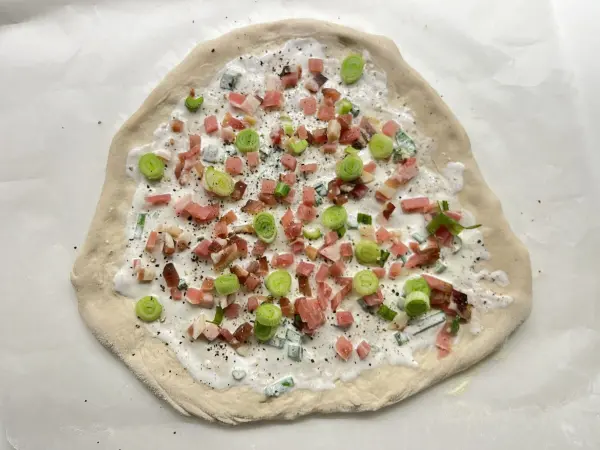 Ein dünner Flammkuchenteig liegt auf Backpapier, bedeckt mit Sauerrahm, Schinken und Frühlingszwiebeln.
