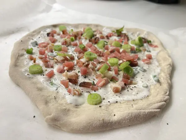 Ein flacher Teig ist mit einer cremigen Sauce und kleinen Stückchen Schinken sowie Frühlingszwiebeln belegt.