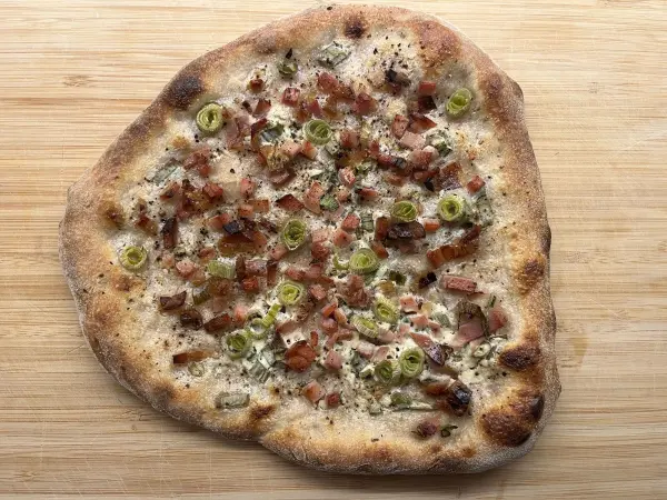Flammkuchen liegt auf einem Holzbrett, belegt mit Schinken und Lauch. Der knusprige Teig sieht appetitlich aus.