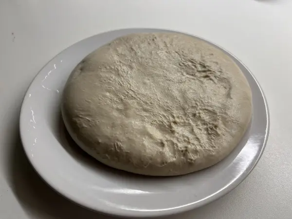 Ein runder, ungekneteter Teig für Flammkuchen liegt auf einem weißen Teller, umgeben von einem neutralen Hintergrund.