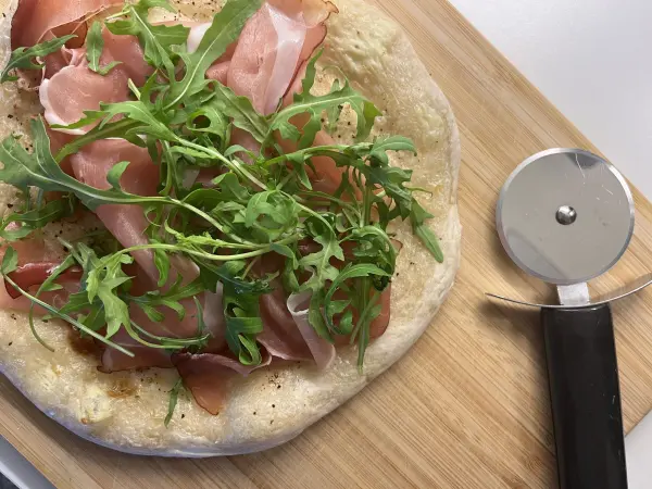 Ein frisch gebackener Flammkuchen liegt auf einem Holzbrett, belegt mit Schinken und Rucola, daneben ein Pizzaschneider.