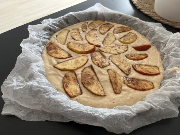 Ein Apfel-Joghurt-Kuchen mit Zimt untermalt, bereit zum Backen, mit frischen Apfelscheiben auf hellem Teig.
