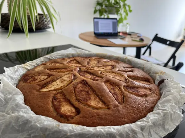 Ein saftiger Apfel-Joghurt-Kuchen mit Zimt ruht auf einem Tisch in einer modernen Küche, umgeben von Pflanzen und einem Laptop.