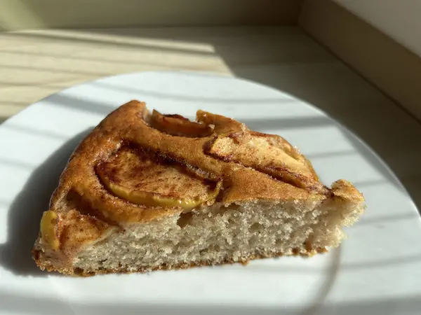 Ein Stück saftiger Apfel-Joghurt-Kuchen mit Zimt liegt auf einem weißen Teller und ist von sanftem Licht umgeben.