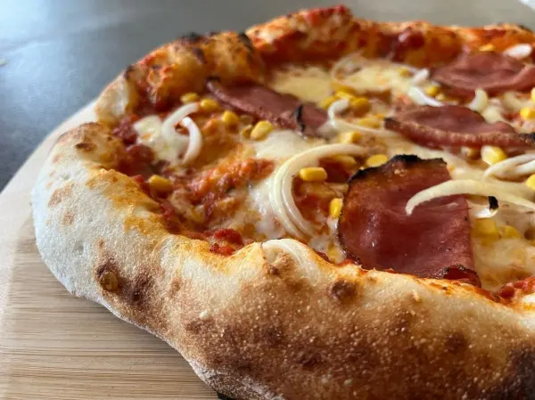 Eine frisch gebackene Pizza mit Schinken, Käse und Mais liegt auf einem Holzbrett und wartet darauf, serviert zu werden.