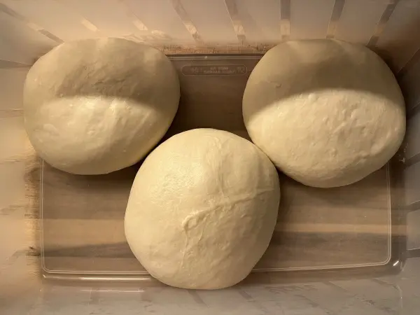 Drei glatte Teigkugeln ruhen in einem durchsichtigen Behälter, bereit zum Ausrollen für die Pizza.