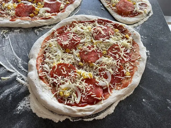 Eine frisch belegte Pizza liegt auf der Arbeitsfläche, überzogen mit Käse, Salami, Zwiebeln und Mais.