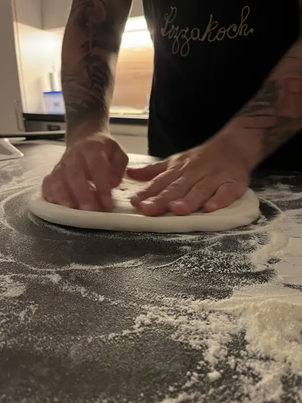 Hände formen einen Pizzateig auf einer mit Mehl bestäubten Arbeitsfläche in einer modernen Küche.