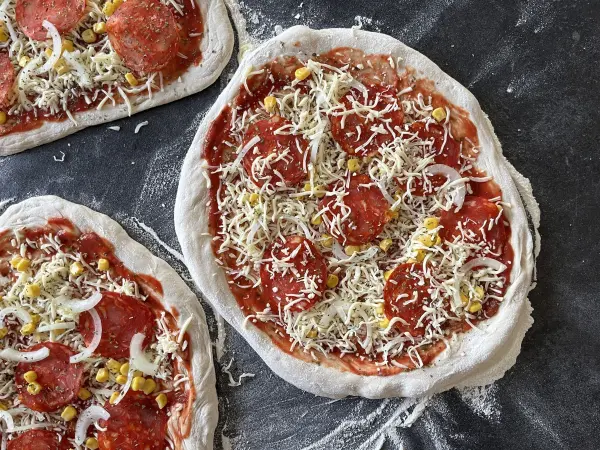 Drei Pizzateigflächen mit Tomatensauce, Käse und Belag liegen auf einer sauberen, bemehlten Fläche zum Backen bereit.