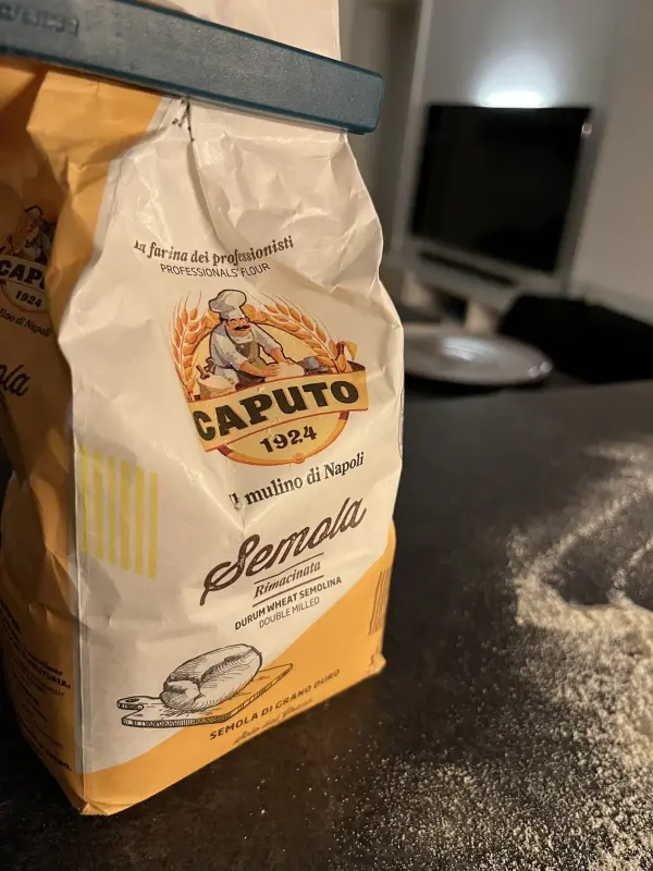 Eine Packung Caputo Semola steht auf einer dunklen Arbeitsplatte, umgeben von feinem Mehl, bereit zum Pizzateigmachen.