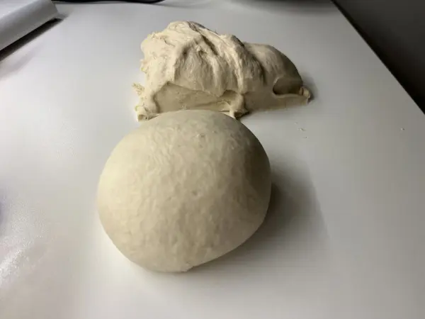 Ein runder Pizzateigball liegt auf einer weißen Oberfläche, während ein Stück frischer Teig daneben ruht.