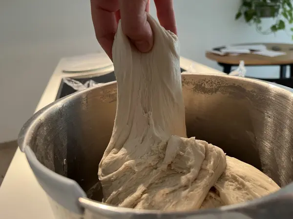 Der Teig wird mit einer Hand aus einer Schüssel gezogen, während der Hintergrund eine helle Küche zeigt.