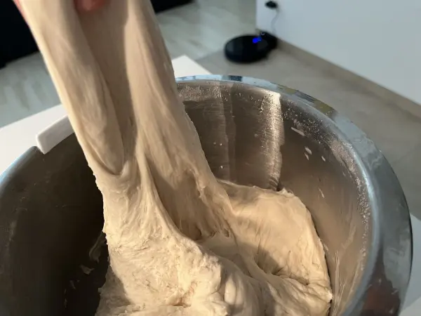 Der elastische Pizza-Teig wird aus einer Schüssel gezogen, während eine moderne Küche im Hintergrund sichtbar ist.