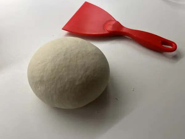 Ein runder Pizzateigball ruht auf einer weißen Arbeitsfläche, daneben liegt ein roter Teigschaber bereit zum Benutzen.