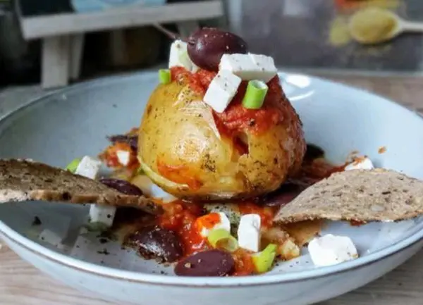 Eine gefüllte Ofenkartoffel steht mittig auf einem Teller, dekoriert mit Tomatensauce, Feta, Oliven und Brot.