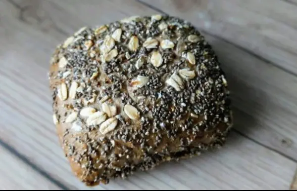 Ein goldbraunes Brötchen mit Haferflocken und Chiasamen liegt auf einem Holztisch, bereit zum Verzehr.