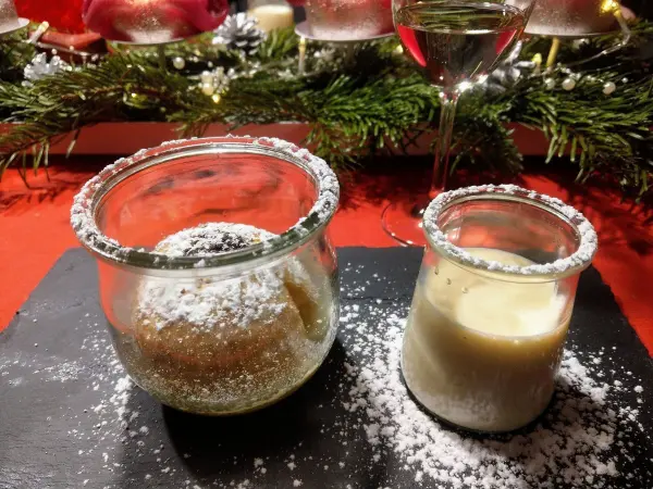 Ein Glas mit einem Bratapfel-Dessert und ein weiteres Glas mit einer cremigen Füllung stehen auf einem schwarzen Teller, umgeben von festlichem Dekor.