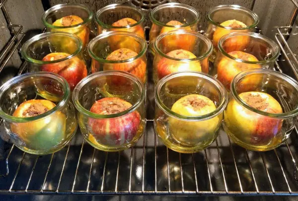 Im Ofen stehen mehrere Gläser mit Bratäpfeln, gefüllt mit Zimt und Zucker, umgeben von duftendem Saft.