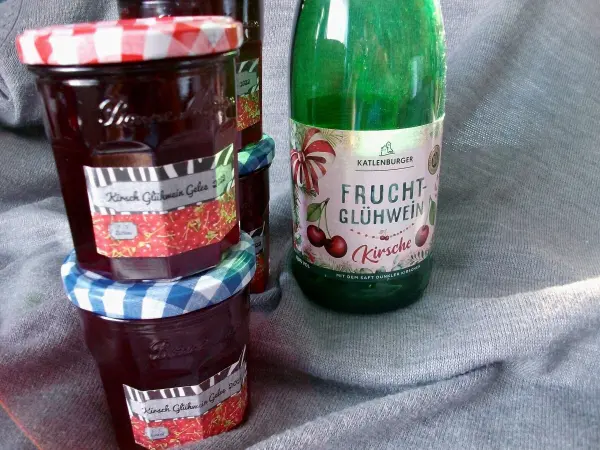 Gläser mit Kirsch-Glühwein-Gelee stehen neben einer Flasche Frucht-Glühwein, alles auf einem grauen Untergrund.