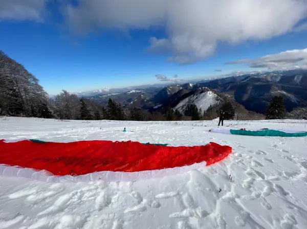Bunte Gleitschirme liegen auf der schneebedeckten Wiese, während jemand sich auf das Gleitschirmfliegen vorbereitet, umgeben von Bergen.