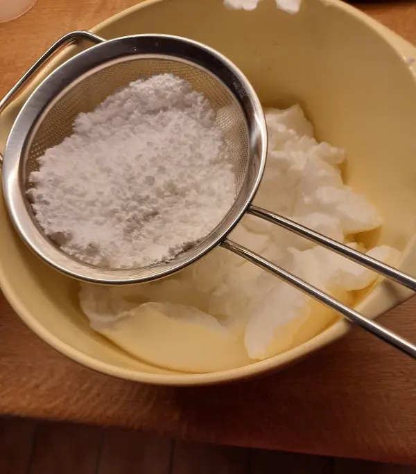 Ein feines Sieb mit Puderzucker wird über eine Schüssel mit fluffigem Eiweiß gehalten, bereit zum Mischen für Haselnussmakronen.