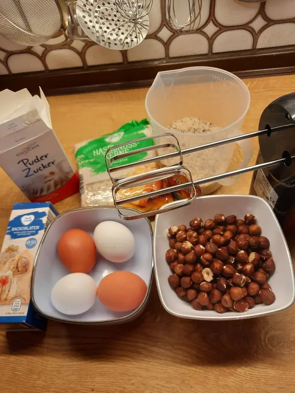 Auf einem Holztisch liegen Zutaten für Haselnussmakronen: Eier, Haselnüsse, Haferflocken und Puderzucker, bereit zum Mischen.