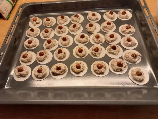 Auf einem Backblech liegt eine große Menge von Haselnussmakronen mit Haferflocken, die auf runden Papptellern platziert sind.