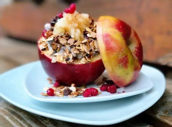 Ein gefüllter Apfel mit Früchtemüsli und Haferflocken steht auf einem weißen Teller in einer natürlichen Umgebung.