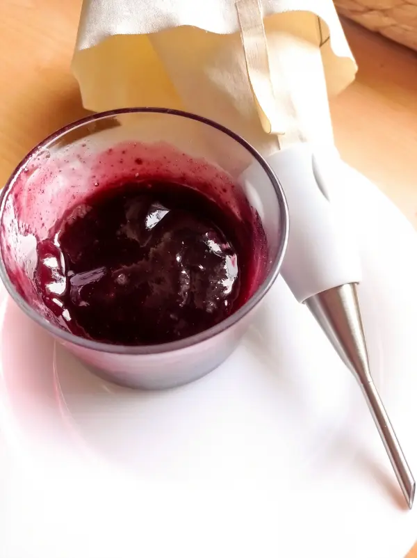 Ein durchsichtiger Becher enthält dunkle Marmelade, neben einem Werkzeug, das zum Füllen von Berliner genutzt wird.