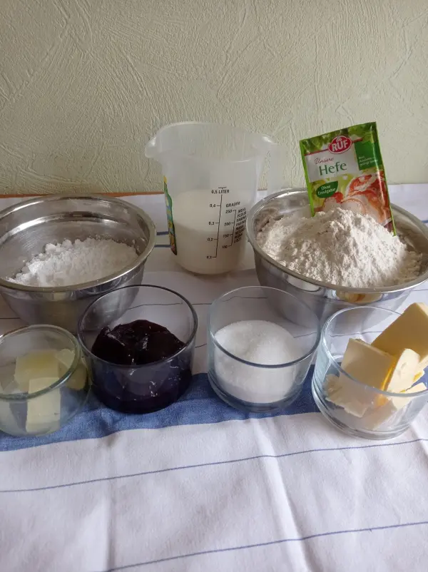 Eine Schüssel mit Mehl steht neben Zutaten wie Butter, Zucker und Marmelade auf einem Tisch, bereit zum Backen von Berliner.