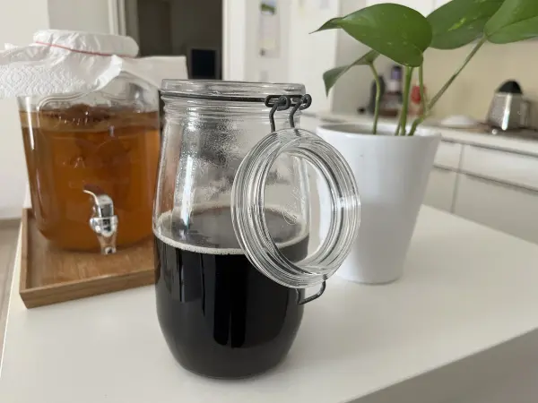 Ein Glas mit dunklem Kaffee steht auf einem Tisch, während im Hintergrund ein anderer Behälter für Kombucha sichtbar ist.