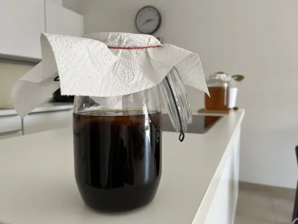 Ein Glas mit dunklem Kaffee steht auf einer Küchenarbeitsfläche, bedeckt mit einem Papiertuch, das schützt.
