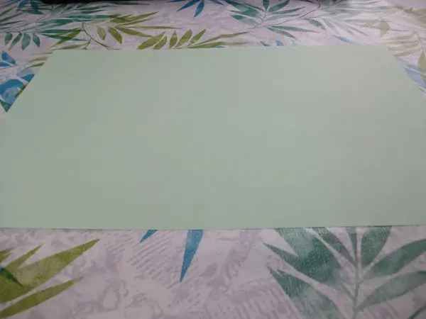 Ein hellgrünes Blatt Papier liegt auf einem blauen und grünen Muster-Tisch. Es ist bereit zum Basteln von Osterhasen.