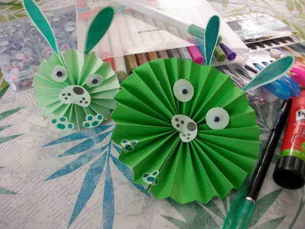 Zwei niedliche Papier-Osterhasen stehen auf einem Tisch, während Bastelmaterialien und Farben im Hintergrund liegen.