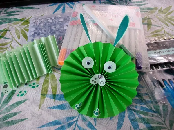 Ein grüner, runder Papier-Osterhase steht auf einem Tisch, umgeben von Bastelmaterialien wie bunten Stiften und Wackelaugen.