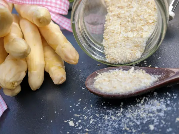 Weißer Spargel liegt neben einem Holzlöffel mit Spargelsalz, während das Salz aus einem offenen Glas auf eine Fläche fällt.