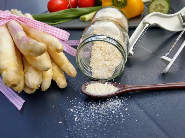 Frischer Spargel liegt mit einem Glas Spargelsalz daneben auf einem Tisch, während etwas Salz auf einen Löffel gefallen ist.