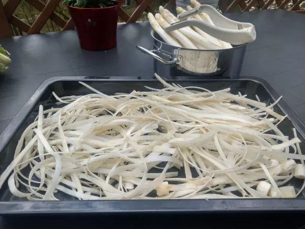 Weiße Spargelreste liegen auf einem Blech, während im Hintergrund frischer Spargel in einem Topf vorbereitet wird.