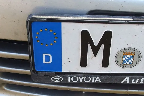 Ein schmutziges Nummernschild mit dem Buchstaben M zeigt, wie Rasierschaum Fliegendreck entfernt. Es ist an einem Toyota befestigt.